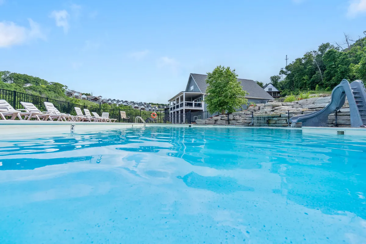 A vibrant exterior shot of the outdoor swimming pool area, showcasing a fun water slide, lounge chairs for sunbathing, and lush greenery surrounding the pool. The building in the background gives a sense of place and the communal aspect of the amenities.