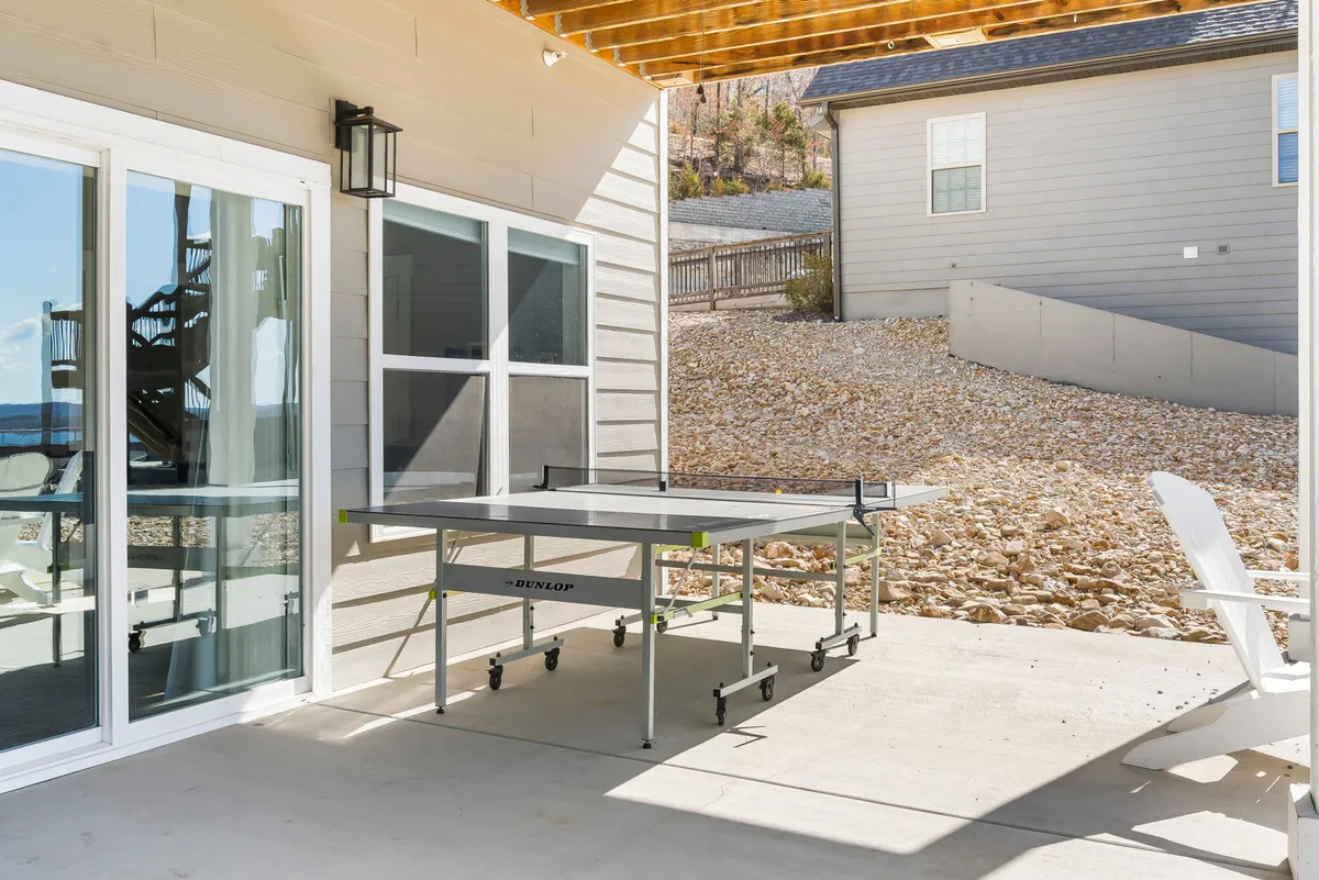 This exterior image features a ping pong table set up on a patio, with sliding glass doors leading inside and comfortable outdoor seating nearby. The background includes lush greenery and a relaxed porch atmosphere, suggesting a seamless blend between indoor and outdoor recreation.