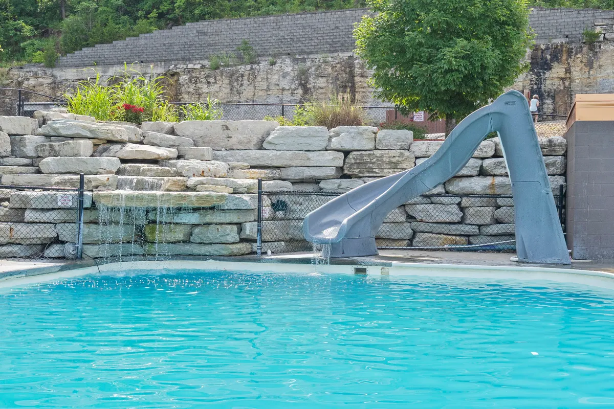 An inviting outdoor swimming pool featuring a curving water slide, rock wall, and a cascading waterfall. Lounge chairs surround the sparkling water, and the scene is filled with sunshine and the vibrant energy of a summer day. The lush greenery and playful design promise fun and relaxation for families and groups.