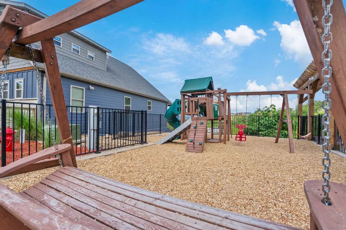 A cheerful playground scene with a swing set and playground structure set on sand, surrounded by a fence. The area is designed for children, with open spaces for play and spots for picnicking or family gatherings.