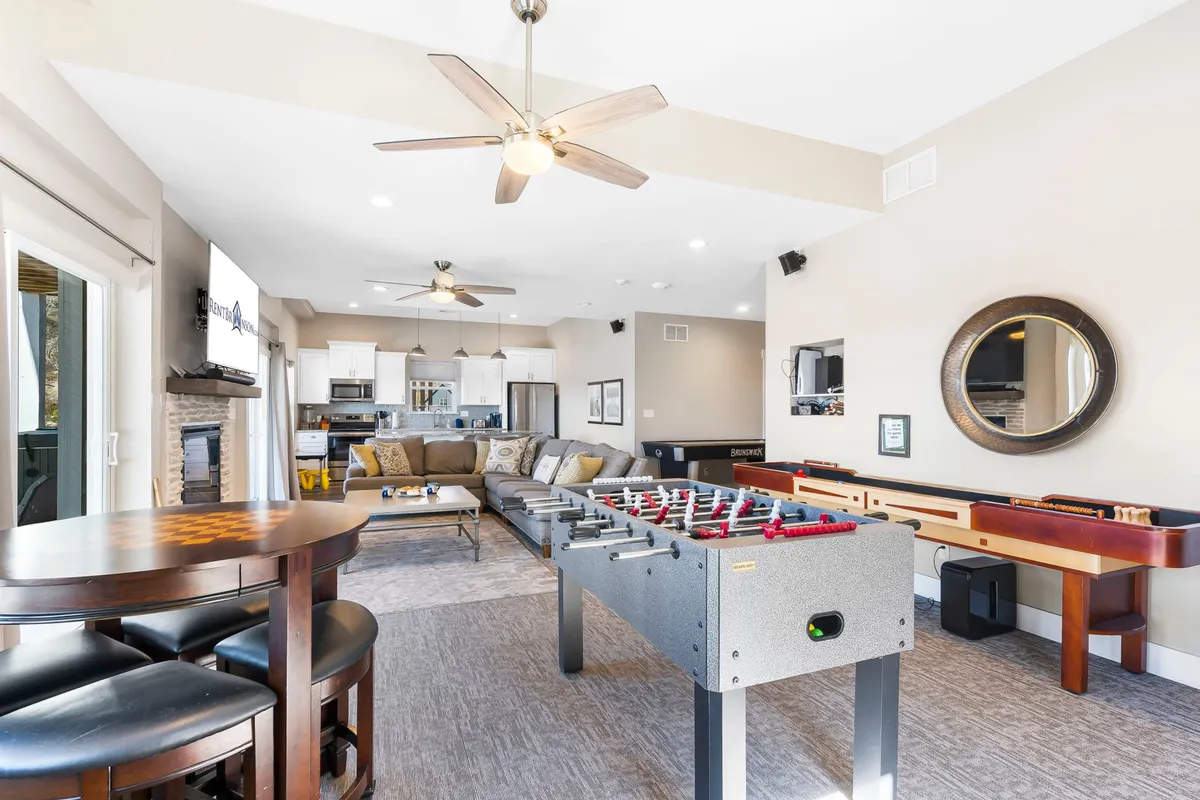 This image features a vibrant game and lounge area, with a foosball table at the forefront, a comfortable seating area, a kitchen space, and a bar area. The setup invites both competitive game play and relaxed socializing, with easy access to snacks and drinks.