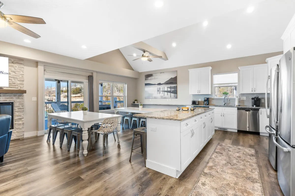 This image showcases a spacious, open-concept kitchen with a large kitchen island and a dining table, seamlessly flowing into a cozy living area with a stone fireplace. The space is filled with natural light from sliding glass doors, and there are glimpses of the scenic lake view beyond. The overall atmosphere is warm and inviting, perfect for group gatherings and shared meals.