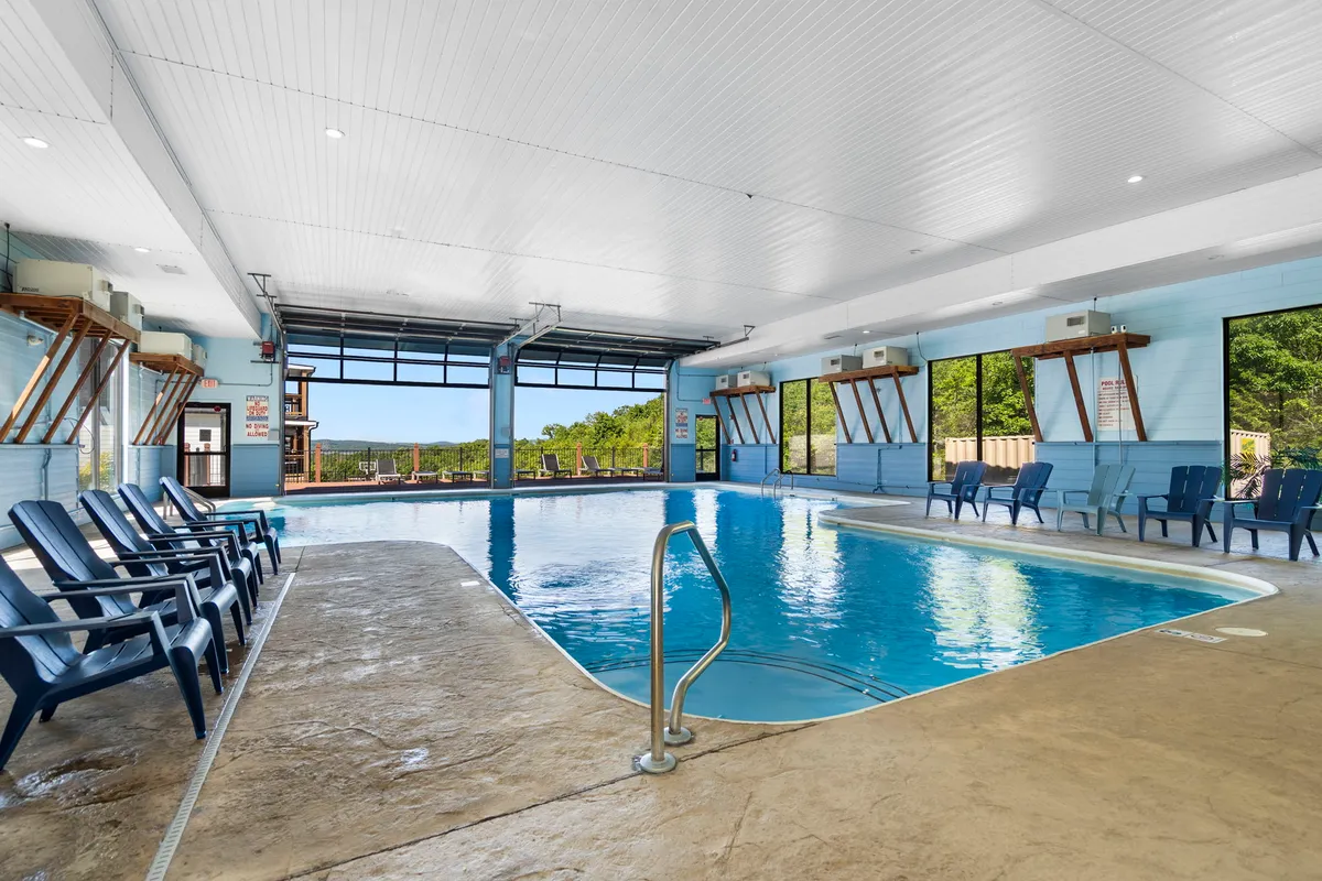 A tranquil indoor swimming pool with large windows letting in natural light and views of the outdoors. Lounge chairs are arrayed poolside, creating a serene and comfortable environment for swimming or relaxing regardless of the weather.