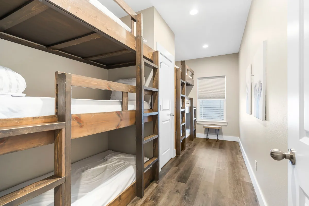 A cozy bunk room featuring multiple bunk beds made of wood, a window with natural light streaming in, and casual decor. The space feels inviting and is clearly designed to accommodate families or groups, encouraging shared experiences and flexible arrangements.
