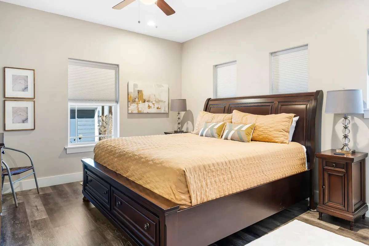 A serene bedroom with a neatly made bed, soft neutral bedding, artwork above the headboard, nightstands on either side, and large windows allowing natural light to fill the space. The overall atmosphere is tranquil and welcoming, suggesting a restful retreat.